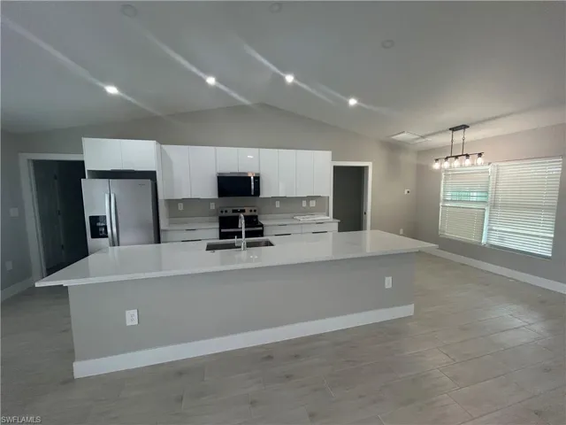 Kitchen featuring stainless steel appliances, white cabinets, a center island with sink, light stone countertops, and vaulted ceiling