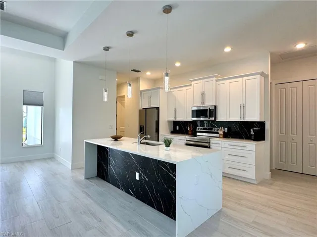 Kitchen with light wood-type flooring, stainless steel appliances, white cabinetry, hanging light fixtures, and an island with sink