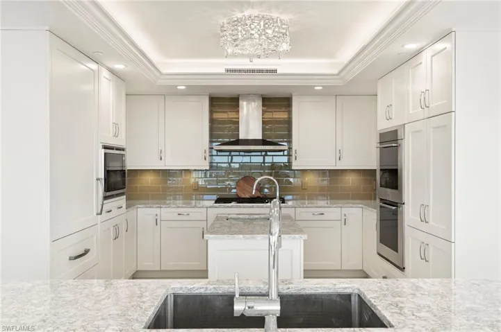 Kitchen featuring white custom cabinetry, a center island with sink, appliances with stainless steel finishes, a tray ceiling, custom backsplash and Custom Cabinet front refrigerator