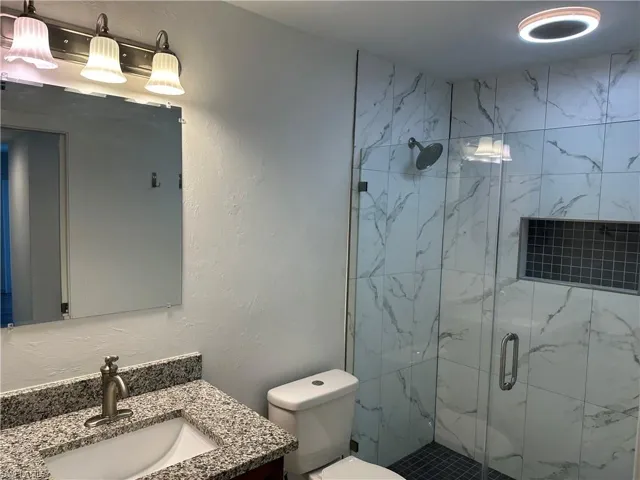 Bathroom featuring a tiled shower with a glass enclosure, a built-in shower niche, a rain showerhead, a vanity with a granite-style countertop, and a brushed nickel faucet