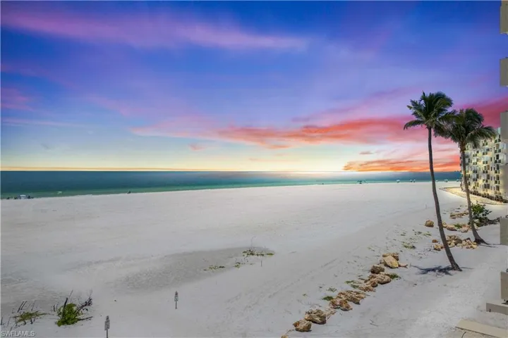 The beach stretches endlessly beneath a sky ablaze with sunset colors—fiery oranges, pinks, and purples melting into the calm Gulf waters.