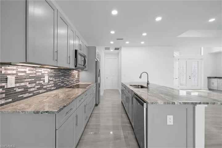 Kitchen with light stone countertops, recessed lighting, a center island with sink, backsplash, and gray cabinetry