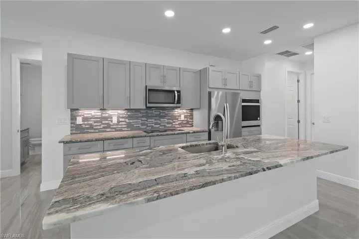 Kitchen with gray cabinetry, light stone countertops, backsplash, stainless steel appliances, and recessed lighting
