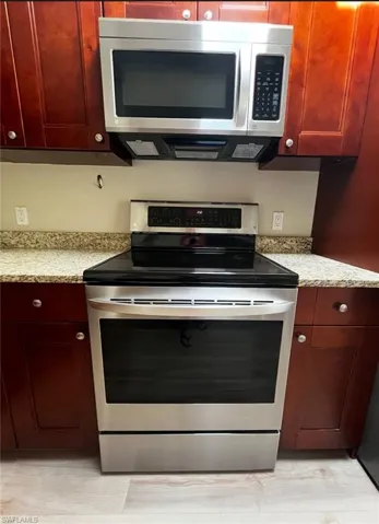 Kitchen with stainless steel appliances, reddish brown cabinets, light wood-style flooring, and light stone counters