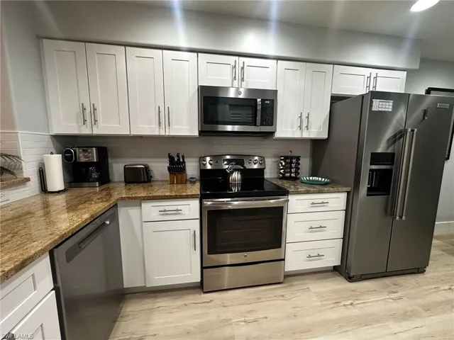 Kitchen with stainless steel appliances, light wood-type flooring, tasteful backsplash, dark stone countertops, and white cabinets