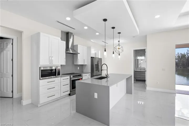 Kitchen with hanging light fixtures, stainless steel appliances, a center island with sink, white cabinets, and light stone counters