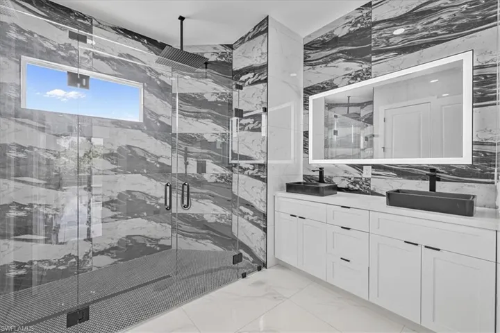 Full bath featuring a marble finish shower, double vanity, light marble finish floors, stone wall, and tile walls