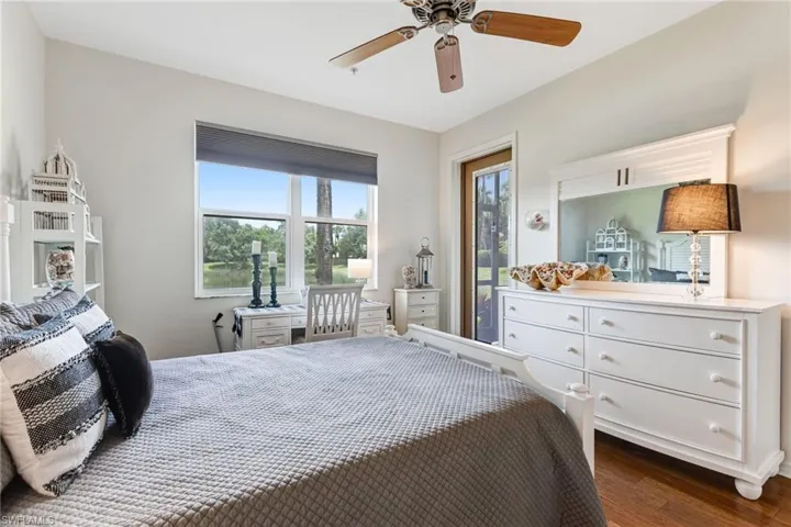 Bright primary bedroom suite featuring hardwood flooring, dual glass doors opening to a private balcony with scenic grounds views, a white painted dresser with ample storage, ceiling fan with remote control, and abundant natural light from large windows