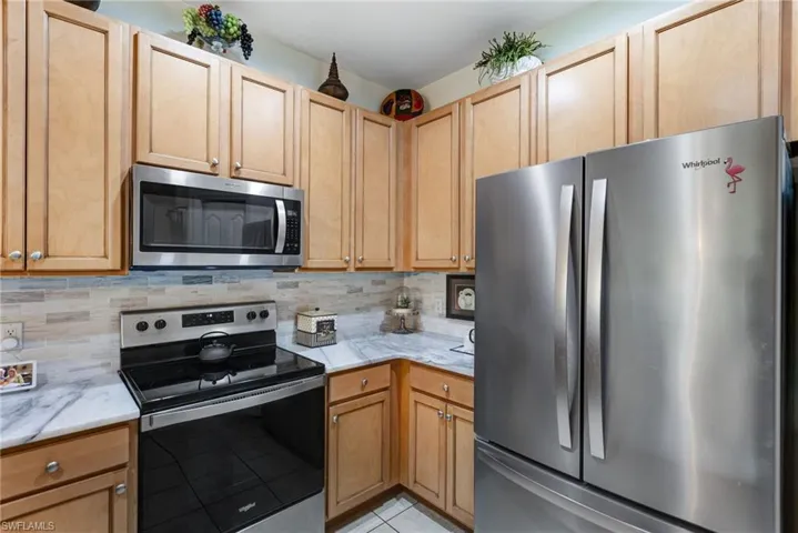 Stainless steel appliances, wood-finish cabinetry, light-toned countertop, tiled backsplash, and tiled flooring
