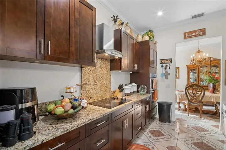 Kitchen with backsplash, dark brown cabinetry, ornamental molding, wall chimney range hood, and light stone counters