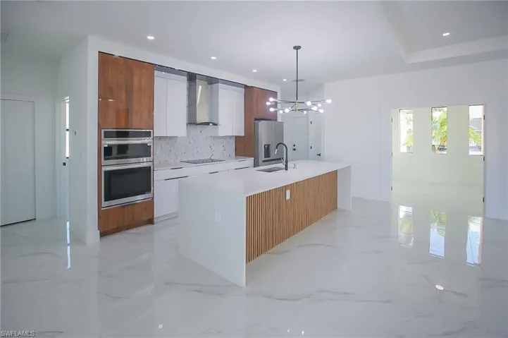 Kitchen featuring white cabinetry, wall chimney exhaust hood, hanging light fixtures, a center island with sink, and appliances with stainless steel finishes