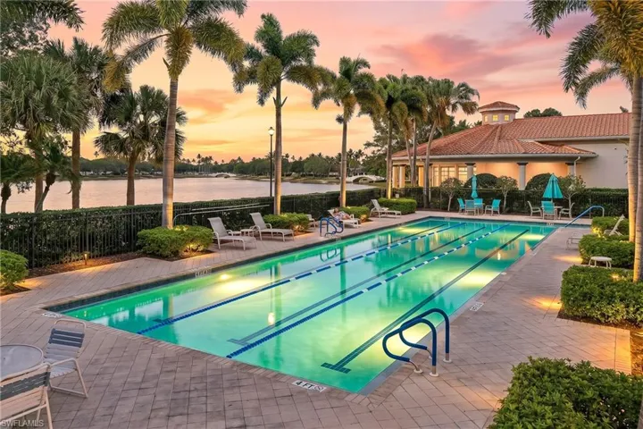 Lap pool with paver decking, surrounded by tropical landscaping, waterfront setting, and a clubhouse with a barrel tile roof