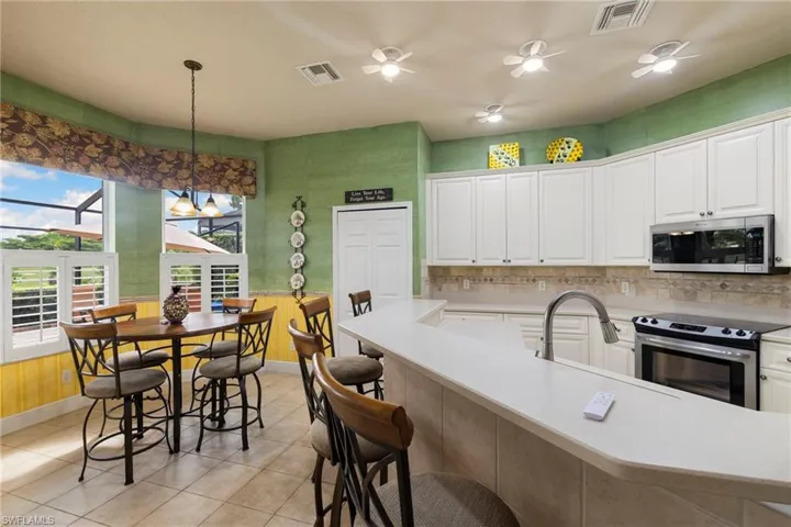 Kitchen featuring appliances with stainless steel finishes, light tile patterned flooring, Eat in table with chairs and bar stools stay, plantation shutters on the lower windows letting in lots sunlight. White cabinets, and light countertops, Fan lights in the kitchen making it cooler to cook on the stove.