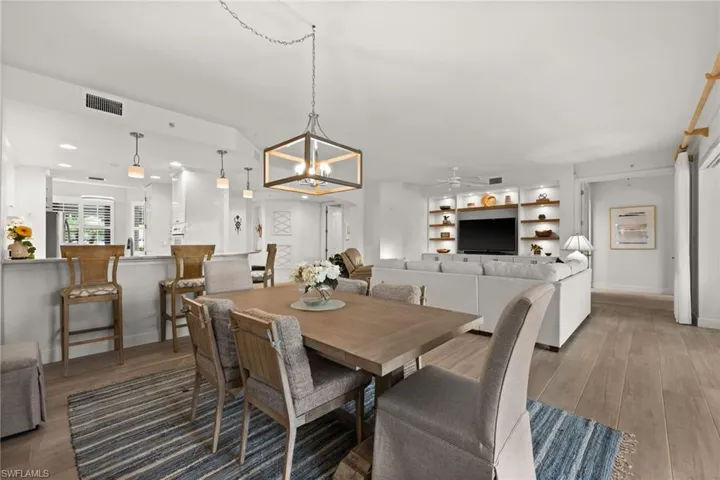 Open-concept living space featuring wood-finish flooring, a built-in media center with shelving, a kitchen island with pendant lighting, and a linear chandelier in the dining area