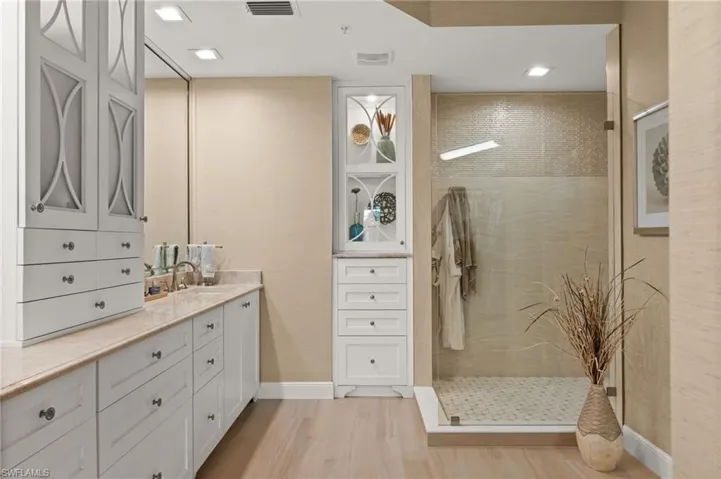 Dual vanity with white cabinetry and stone countertop, glass-enclosed shower with tiled walls, wood-finish flooring, recessed lighting, and built-in storage