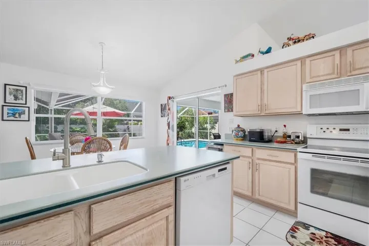 Kitchen with light brown cabinetry, white appliances, vaulted ceiling, light tile patterned floors, and hanging light fixtures