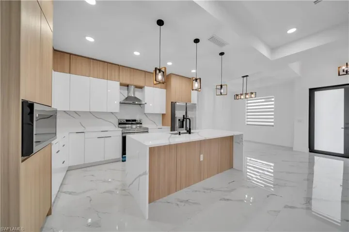 Kitchen with decorative light fixtures, two tone cabinets, stainless steel appliances, light stone countertops, and a kitchen island with sink