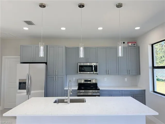 Kitchen with light stone counters, stainless steel appliances, a kitchen island with sink, and pendant lighting