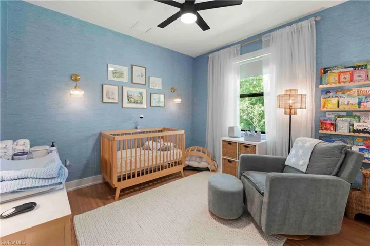Textured blue wall covering, wood-finish flooring, ceiling fan with integrated lighting, wall-mounted light fixtures, and a large window with sheer drapery