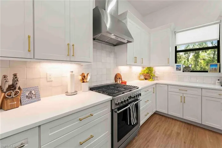 White shaker-style cabinetry with gold-tone hardware, stainless steel range with matching hood, light-toned countertops, and wood-finish flooring