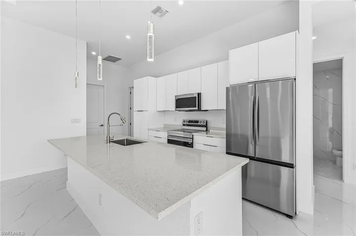 Kitchen featuring stainless steel appliances, pendant lighting, white cabinetry, light stone countertops, and a kitchen island with sink