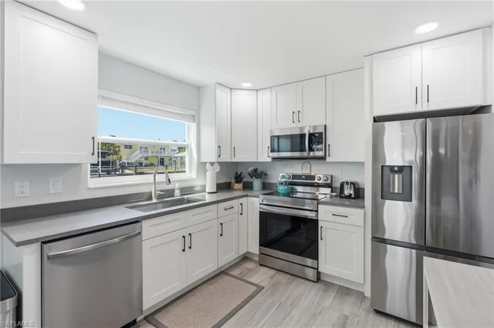 Modern kitchen featuring white shaker cabinetry, stainless steel appliances, gray countertops, a stainless steel sink with a gooseneck faucet, and light wood-finish flooring - Virtually Edited Image