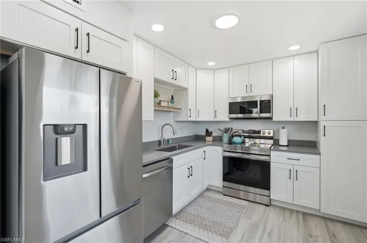 Modern kitchen featuring white shaker cabinetry with matte black hardware, stainless steel appliances, gray countertops, and wood-finish flooring - Virtually Edited Image