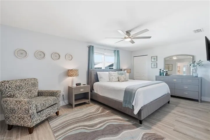 Spacious bedroom featuring wood-finish flooring, a contemporary ceiling fan, window with light-filtering curtains, and a built-in closet with bi-fold doors - Virtually Edited Image