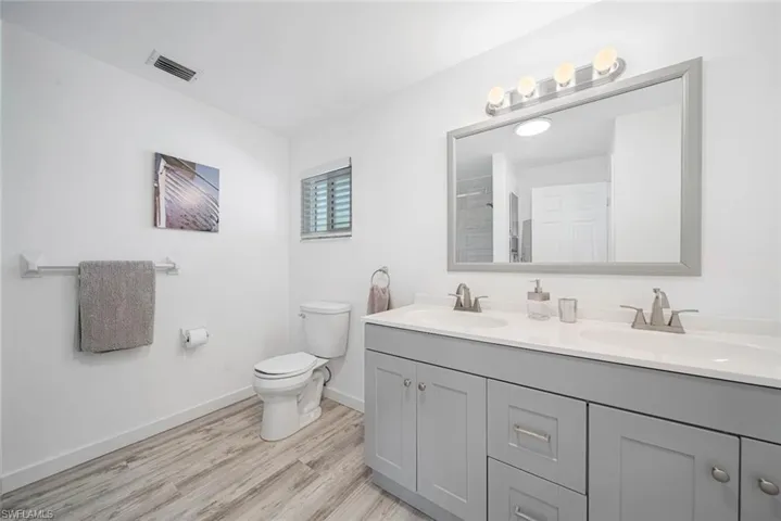 Dual-sink vanity with gray shaker-style cabinetry, white countertop, large framed wall mirror, four-light fixture, and wood-finish flooring