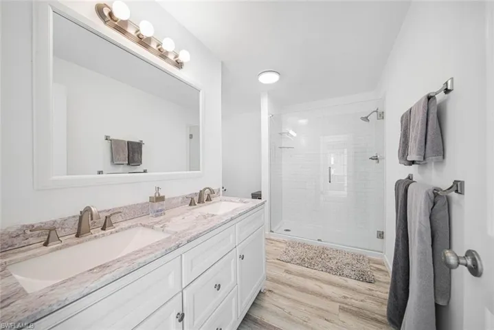 Dual vanity featuring white cabinetry, stone-finish countertops, undermount sinks, and a framed mirror with integrated four-light fixture, paired with a glass-enclosed shower featuring white tiled walls and wood-finish flooring