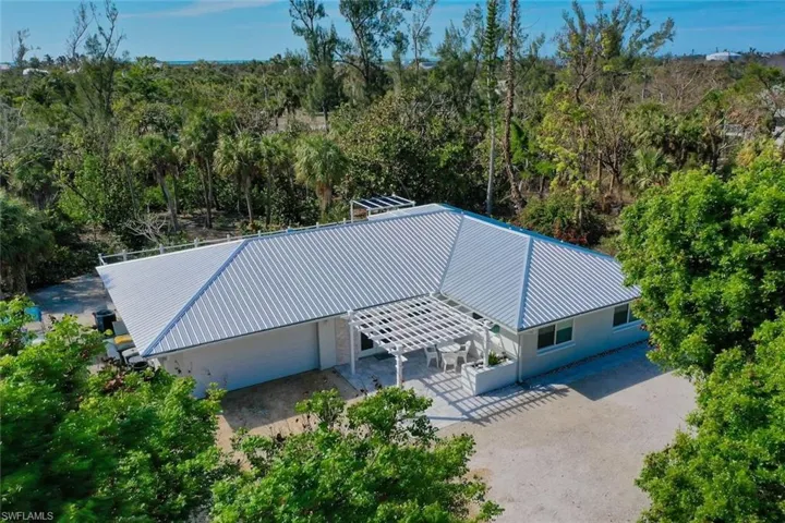 Single-story residence featuring a standing-seam metal roof, white exterior cladding, attached pergola, and a gravel driveway, surrounded by mature trees