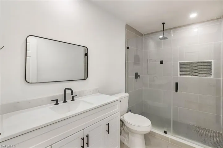 Bathroom featuring a white vanity with a rectangular undermount sink and matte black faucet, a framed rectangular mirror, a toilet, and a glass-enclosed shower with gray tiled walls and a built-in niche
