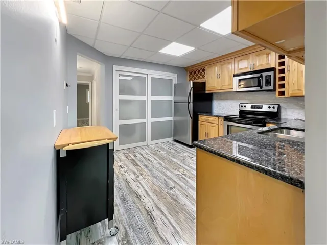 Kitchen featuring light wood-type flooring, tasteful backsplash, stainless steel appliances, a drop ceiling, and dark stone counters
