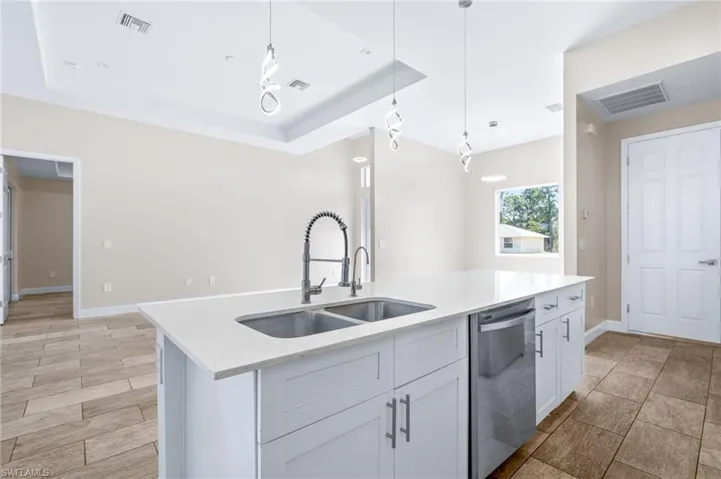 Kitchen featuring a raised ceiling, stainless steel dishwasher, light stone counters, decorative light fixtures, and an island with sink