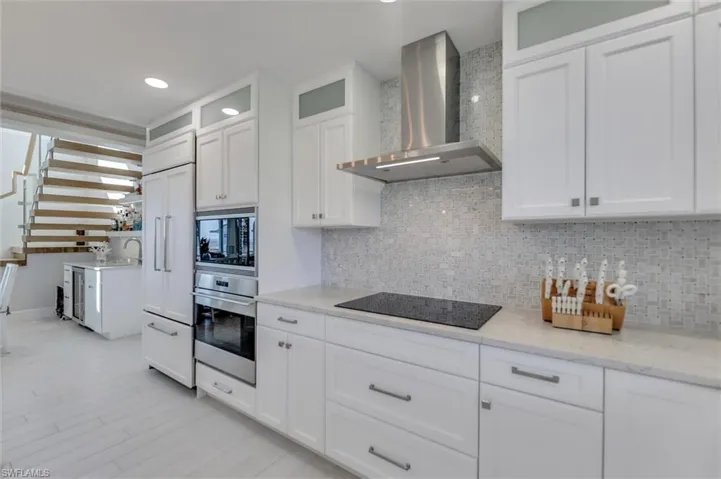 Kitchen with white cabinetry, wall chimney range hood, black appliances, glass insert cabinets, and light stone counters
