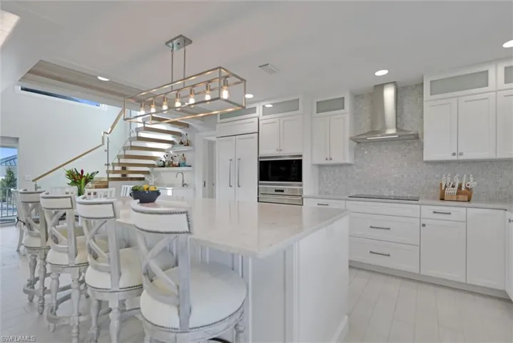 Kitchen featuring white cabinetry, light stone counters, glass insert cabinets, wall chimney range hood, and recessed lighting