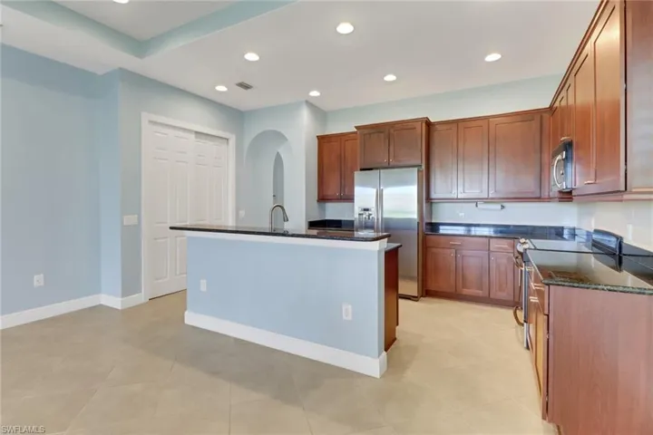 Kitchen featuring recessed lighting, stainless steel appliances, stone counters, a center island with sink, and brown cabinetry
