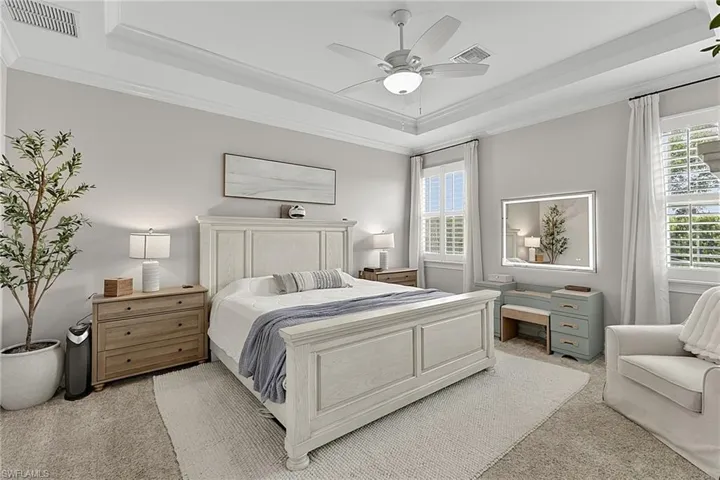 Spacious bedroom featuring a tray ceiling, ceiling fan with integrated lighting, and two windows with plantation shutters