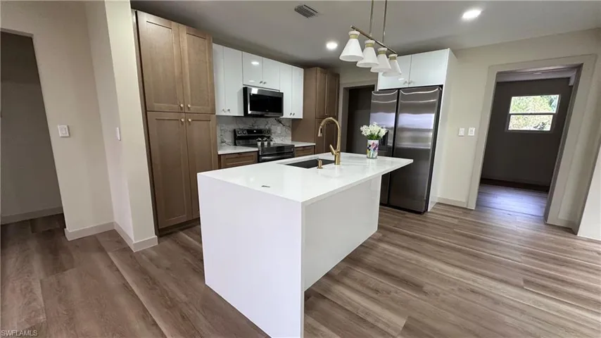 Kitchen featuring stainless steel appliances, light stone countertops, an island with sink, hanging light fixtures, and light wood-style floors