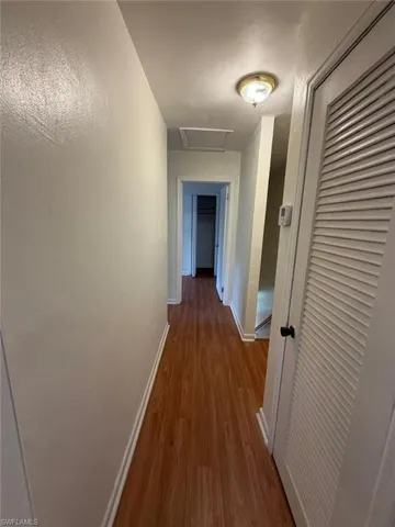 Wood-finish flooring throughout the hallway, featuring white baseboards, a flush-mount ceiling light fixture, and a white louvered door