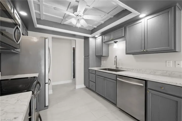 Kitchen featuring stainless steel appliances, gray cabinetry, ceiling fan, coffered ceiling, and light stone countertops