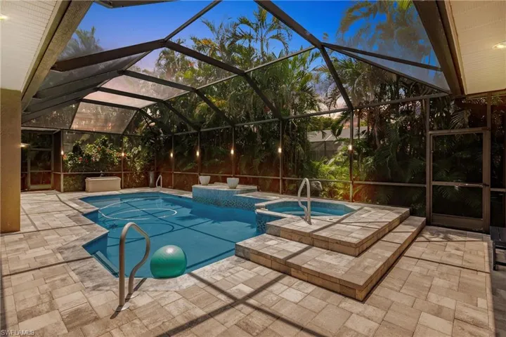 Pool at dusk with an in ground hot tub, glass enclosure, and a patio area