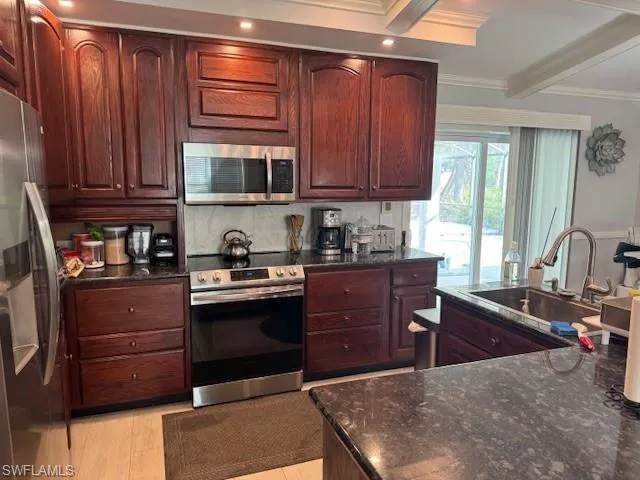 Kitchen with beamed ceiling, stainless steel appliances, dark stone counters, ornamental molding, and sink