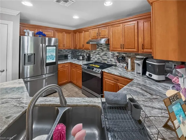 Kitchen with stainless steel appliances, wood finish cabinetry, light tile patterned floors, tasteful backsplash, and light stone counters