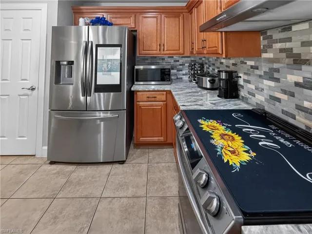 Kitchen featuring stainless steel appliances, wood finish cabinets, light tile patterned flooring, backsplash, and light stone countertops