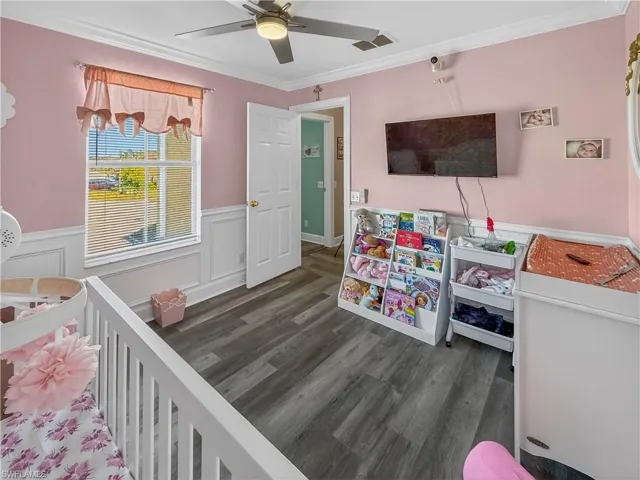 Bedroom with dark wood finished floors, ornamental molding, a nursery area, a wainscoted wall, and a decorative wall