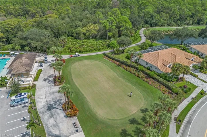 Aerial perspective featuring an expansive manicured lawn, paved parking area, tile-roofed structures, palm tree landscaping, and proximity to a water feature