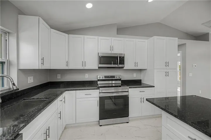 Kitchen featuring stainless steel appliances, dark stone countertops, white cabinets, lofted ceiling, and light marble finish flooring