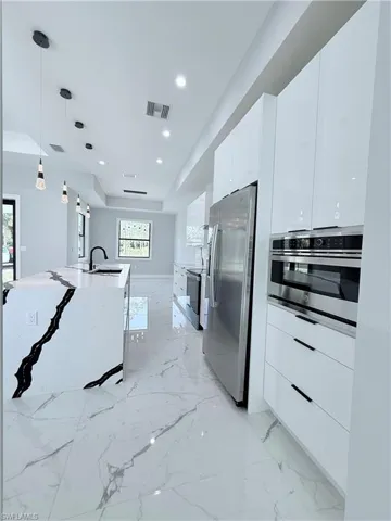 Kitchen featuring a kitchen island with sink, hanging light fixtures, light stone counters, white cabinetry, and stainless steel appliances