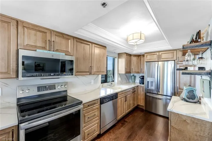 Kitchen with appliances with stainless steel finishes, light stone counters, dark wood-style flooring, crown molding, and a tray ceiling